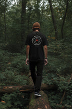 Person walking on a log in a forest wearing a black t-shirt with a logo.